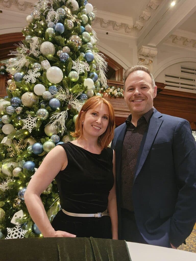 A man and a woman in formal wear pose before a Christmas tree.