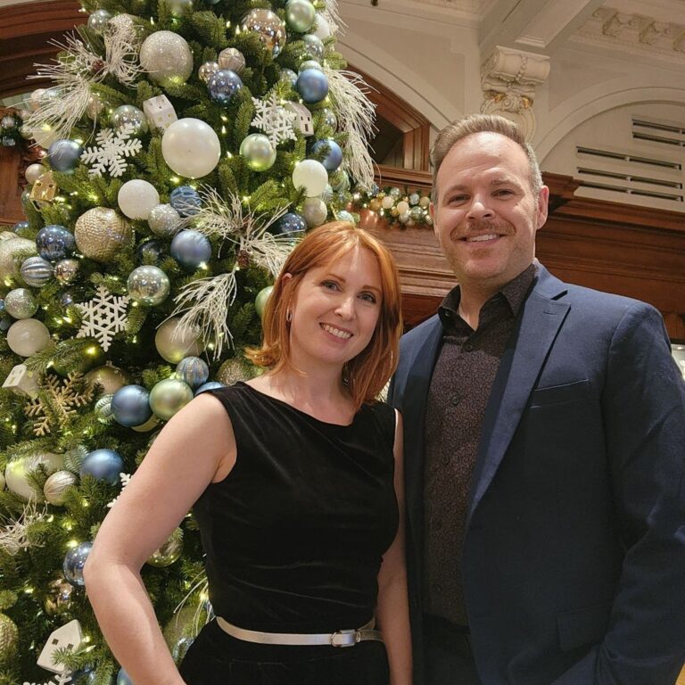 A man and a woman in formal wear pose before a Christmas tree.