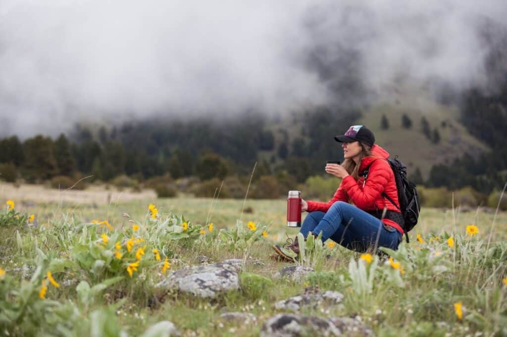 A woman in a red jacket sits in a mountain field to sip a warm drink from a thermos.