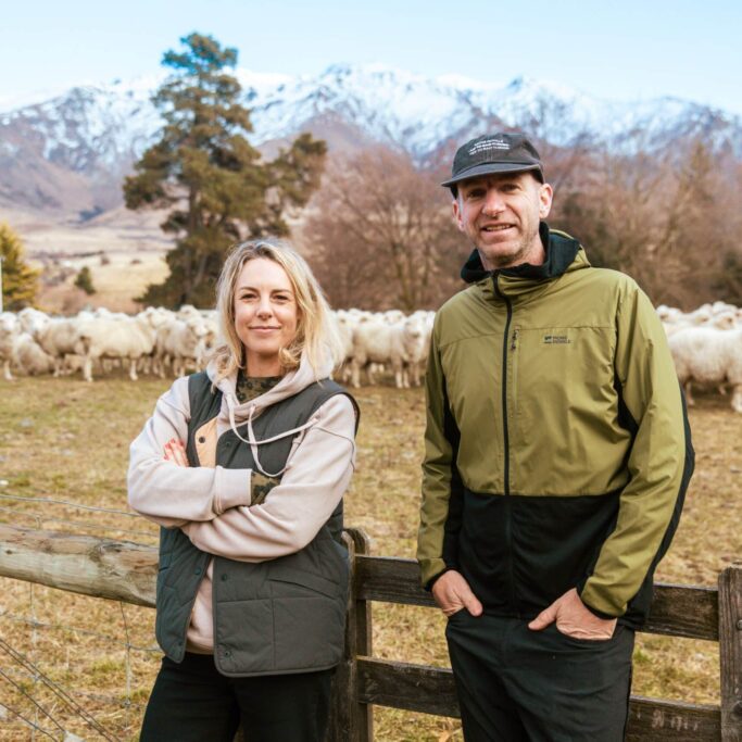 Hamish & Hannah, founders of Mons Royale stand on a farm in front of a field of sheep.