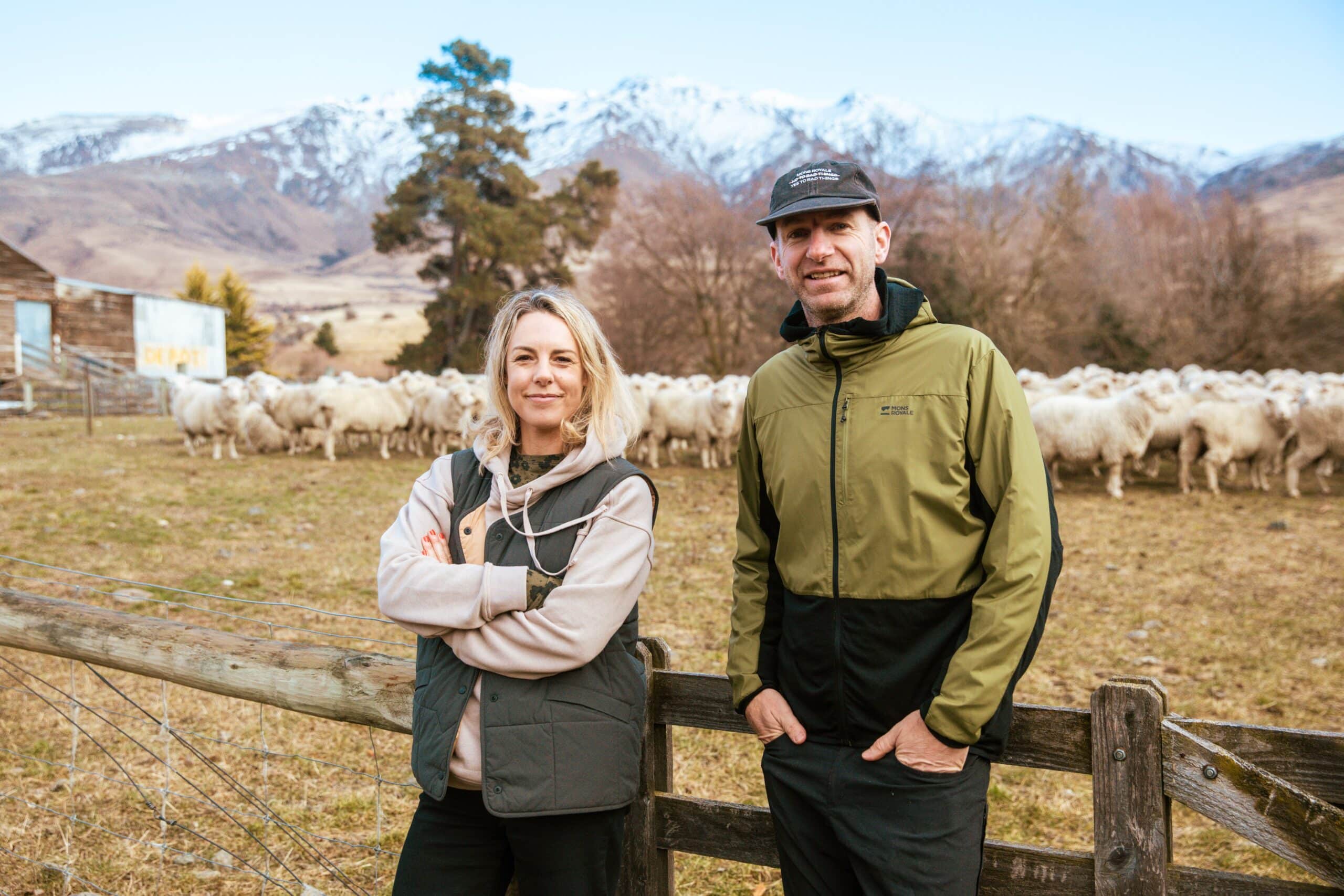 Hamish & Hannah, founders of Mons Royale stand on a farm in front of a field of sheep.