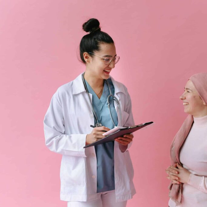 A woman wearing scrubs and a lab coat speaks to another woman and takes notes on a clipboard. Behind them is a pink background.