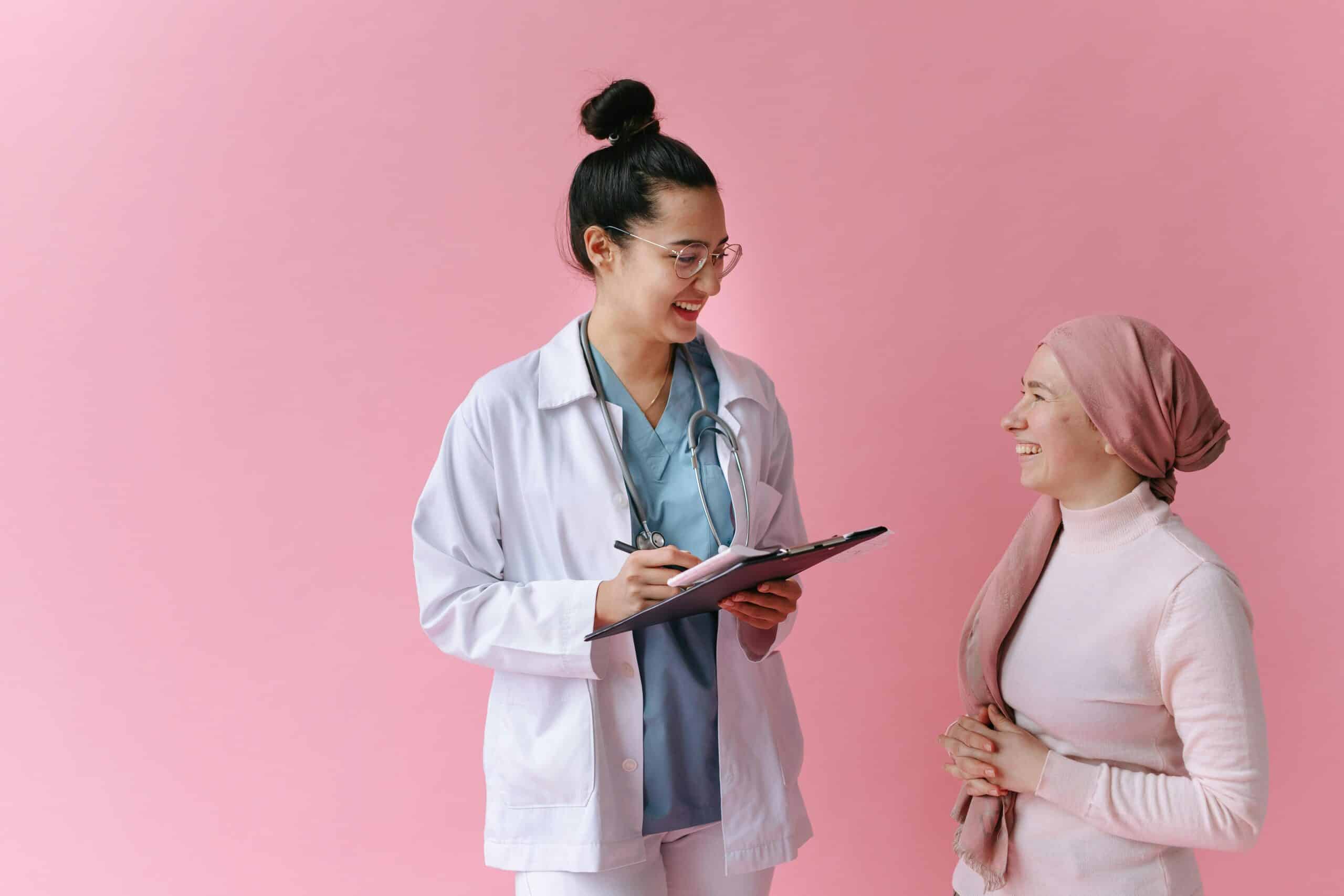 A woman wearing scrubs and a lab coat speaks to another woman and takes notes on a clipboard. Behind them is a pink background.