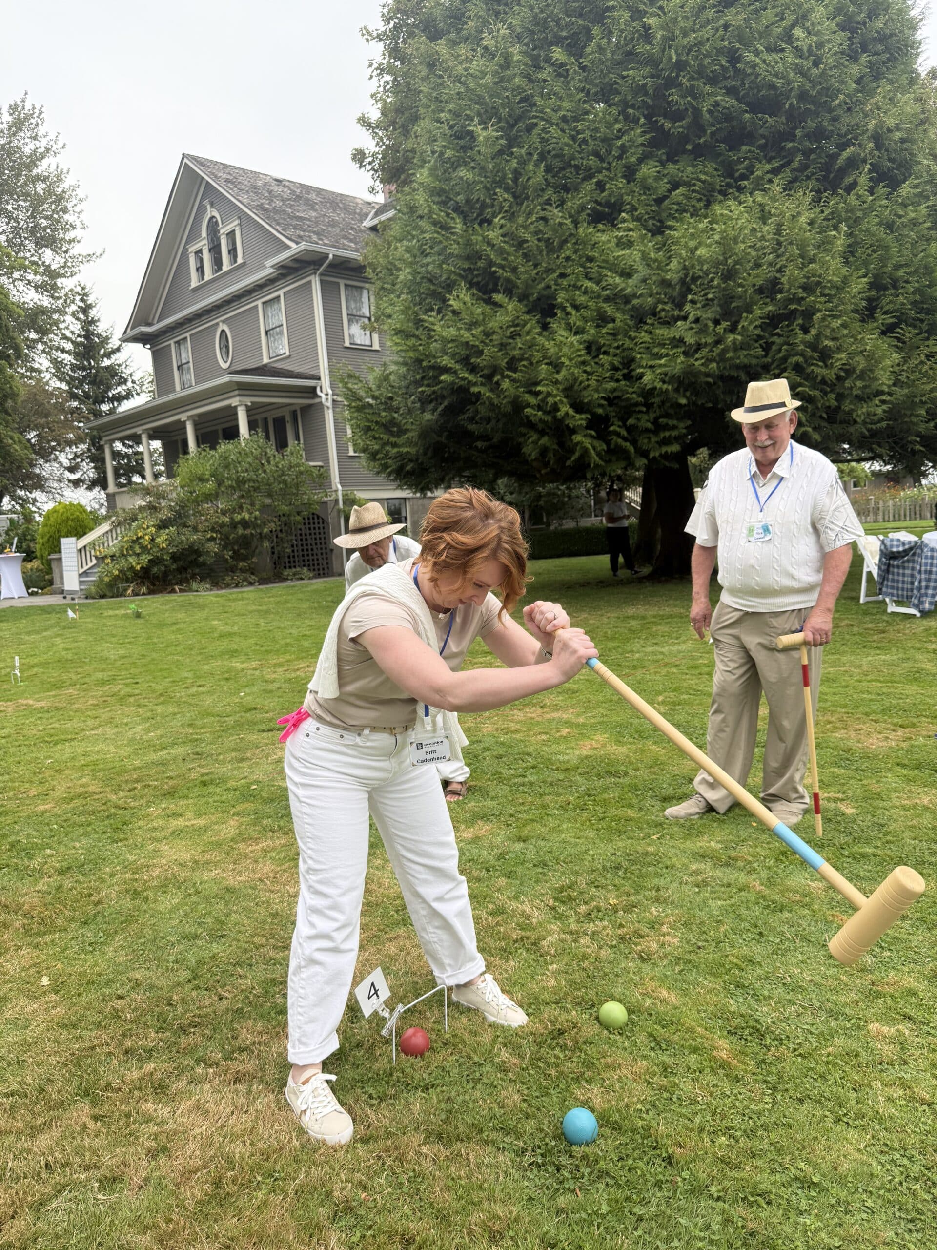 A woman hits a blue croquet ball on a lawn in front of a house. 