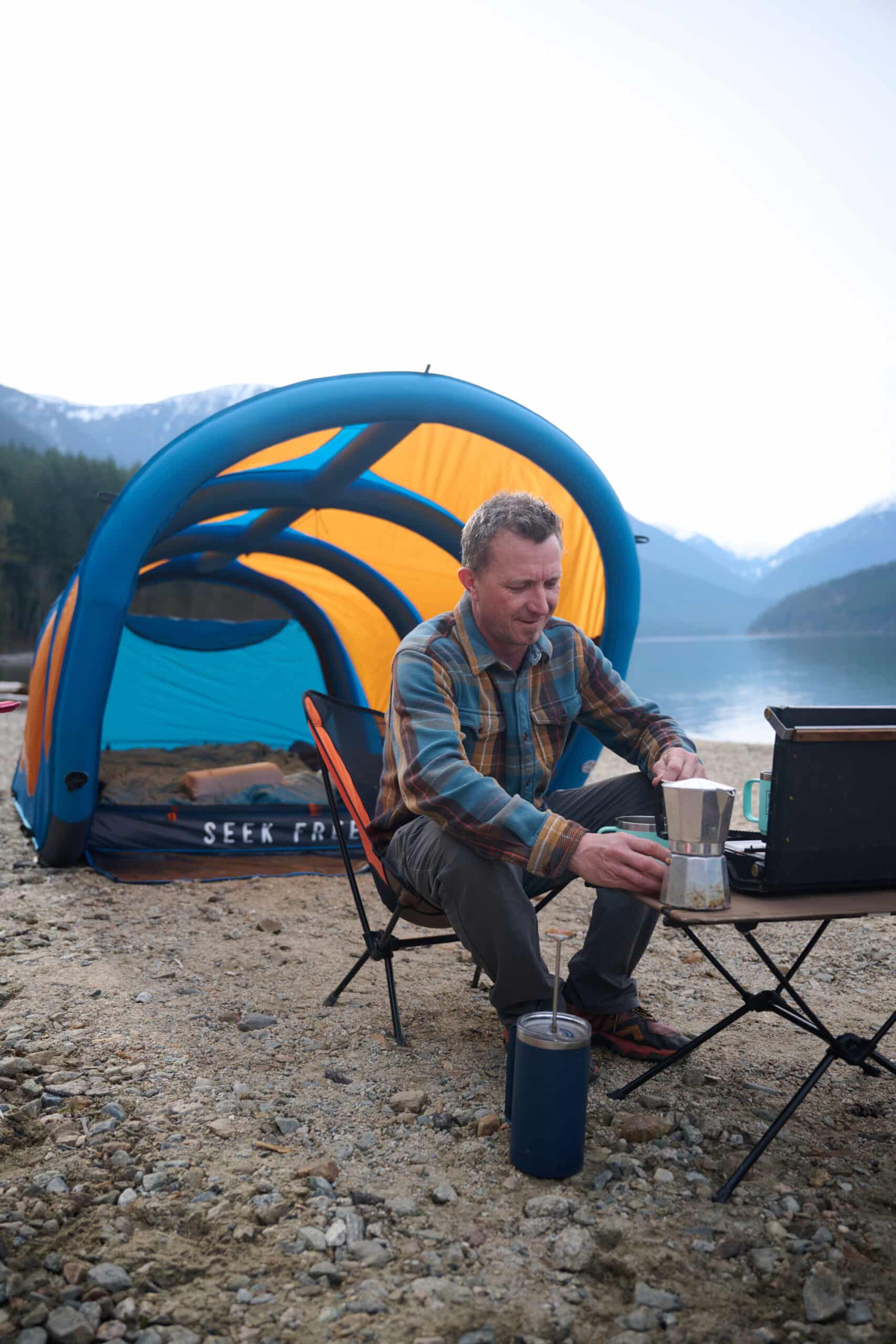 A man sits in a camping chair and makes coffee in front of an inflatable tent