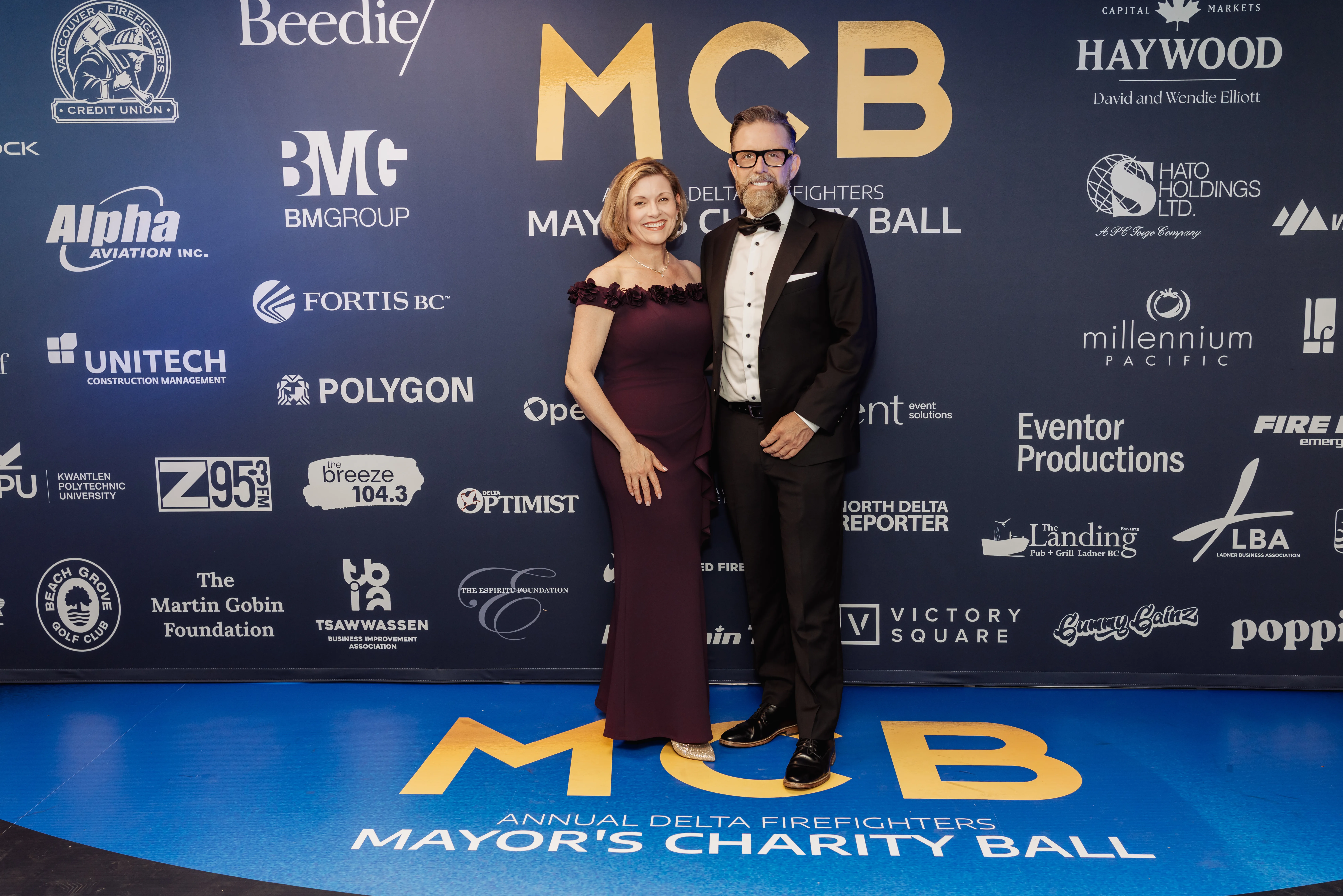 Evolution Group Of Companies Founders, Gwen and Ben Martin attend the Annual Delta Firefighters Mayor’s Charity Ball. The stand in front of a sign that says MCB Mayor's Charity Ball.